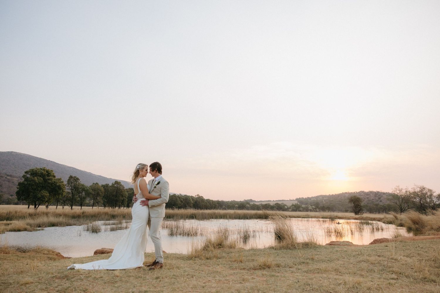 Wedding couple posing in scenic field with water and mountains in background at sunset.
