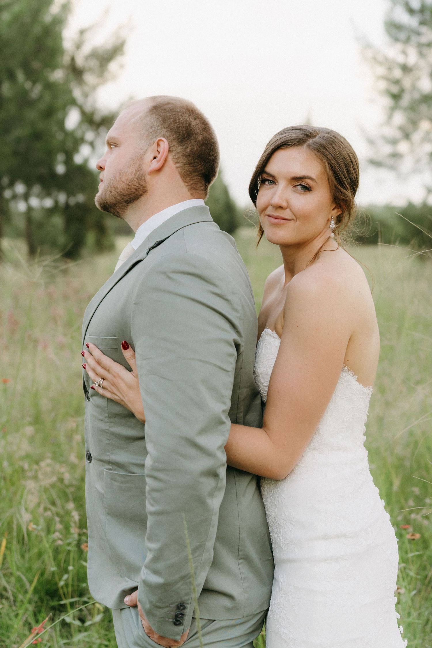 A romantic wedding photo of a couple in a meadow, with the bride in white embracing the groom in a gray suit from behind.