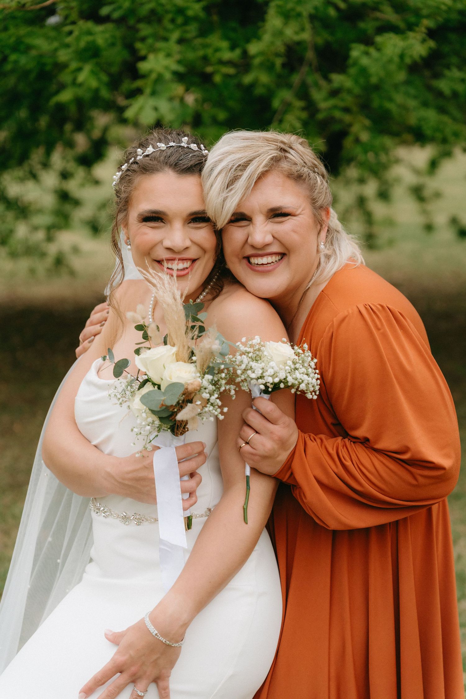 Wedding party in white and burnt orange dresses pose playfully outdoors under green trees.