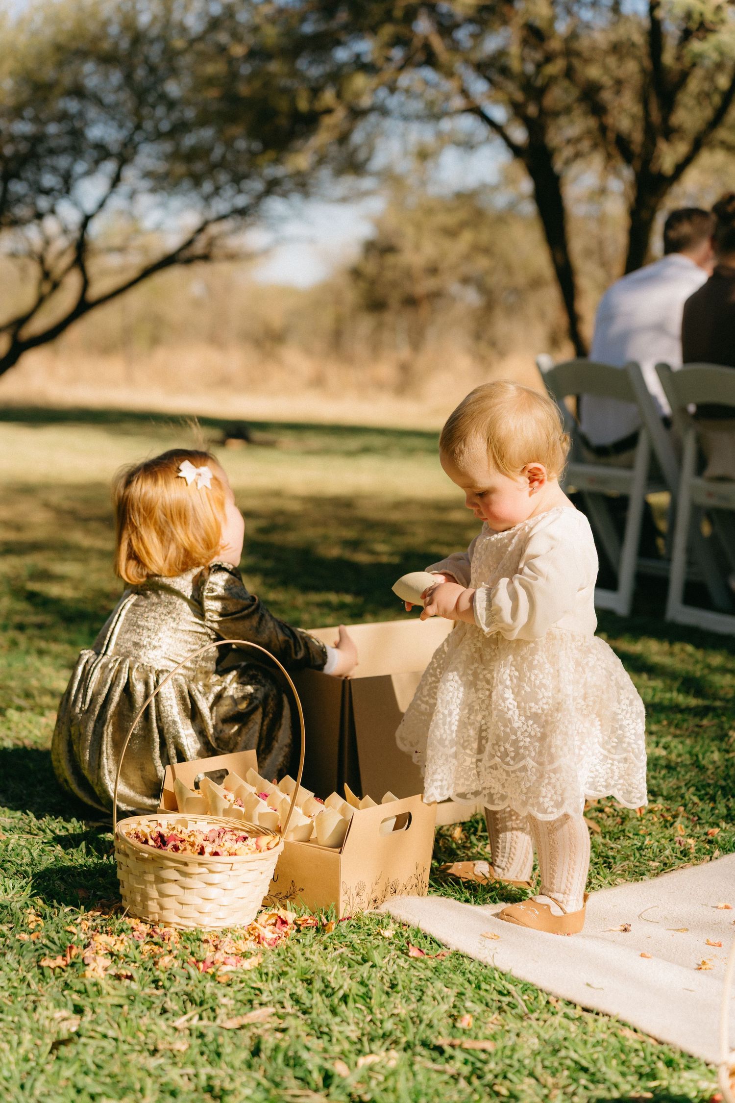 Young children in white outfits play with baskets on a grassy lawn during a wedding.