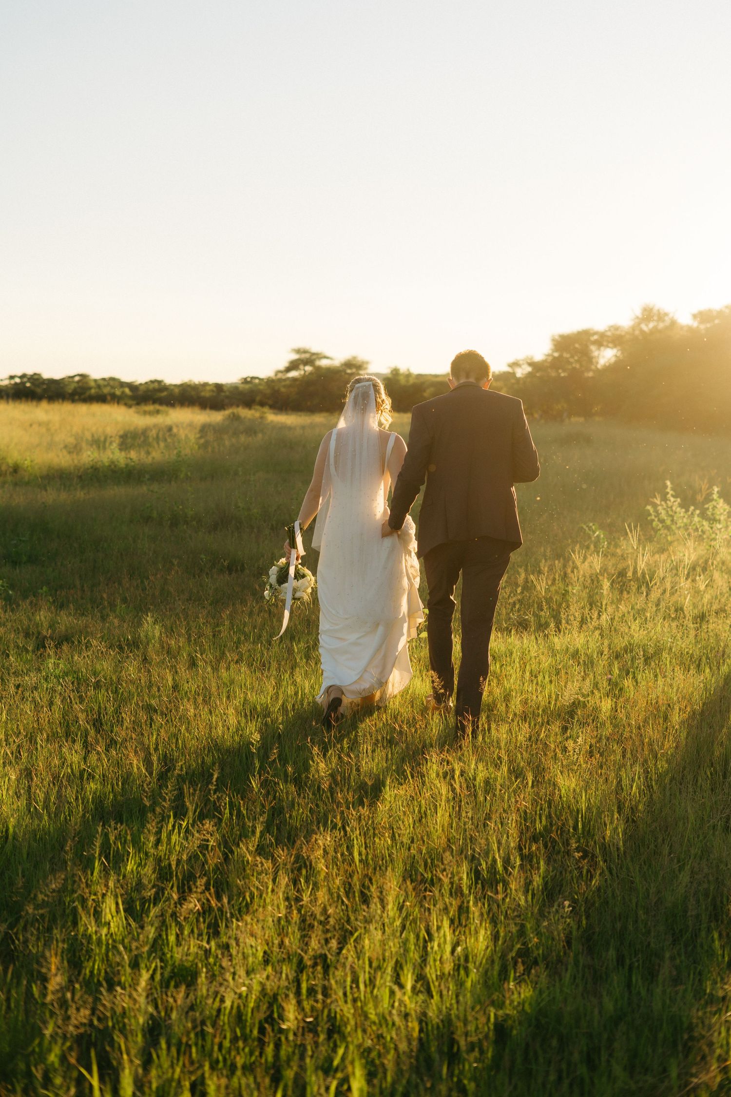 A couple walks hand in hand through a golden field at sunset on their wedding day.