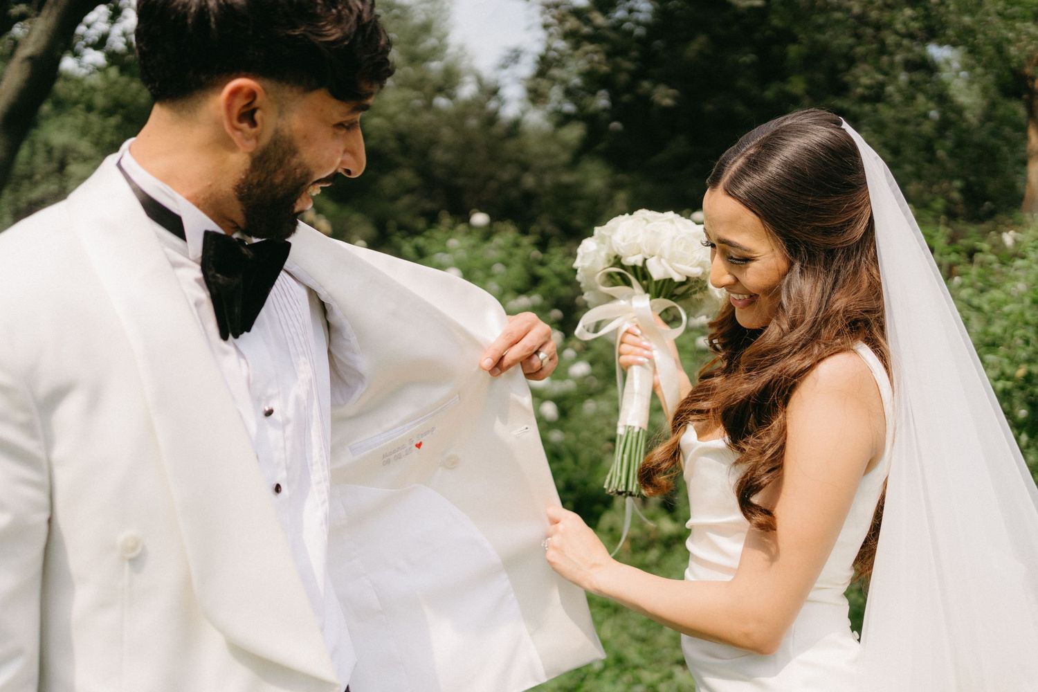 A joyful wedding couple shares a candid moment with their small white fluffy dog during an outdoor photoshoot.