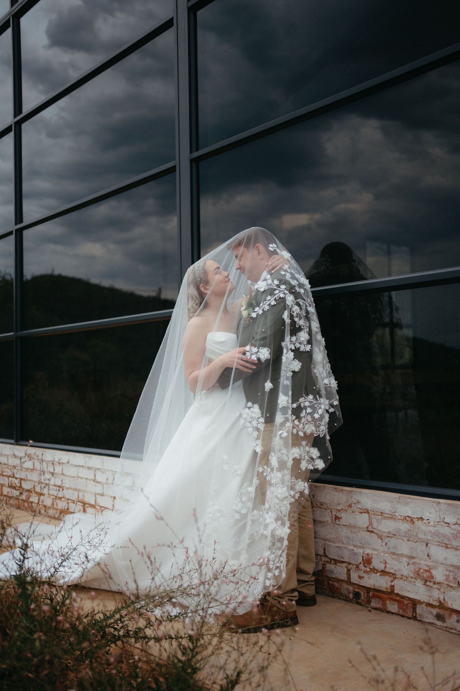 A dramatic wedding photo against dark storm clouds shows an intricate lace veil blowing in the wind by large glass windows.