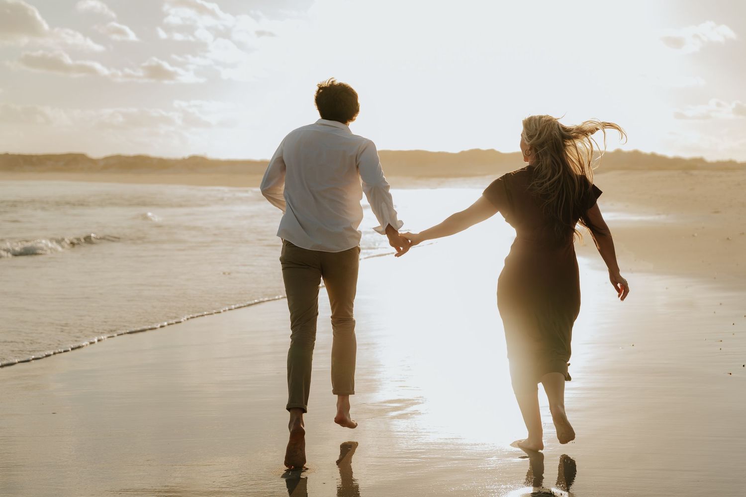 Couple holding hands running barefoot on a sandy beach at golden sunset with ocean waves in the background.