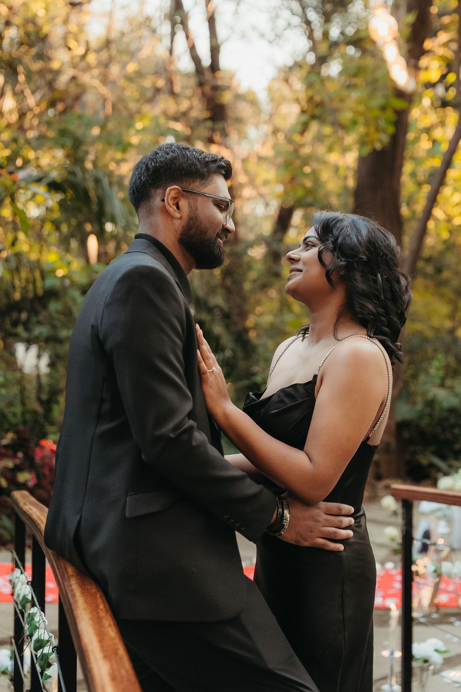Couple in matching black outfits sharing a romantic moment on a balcony surrounded by lush green trees.