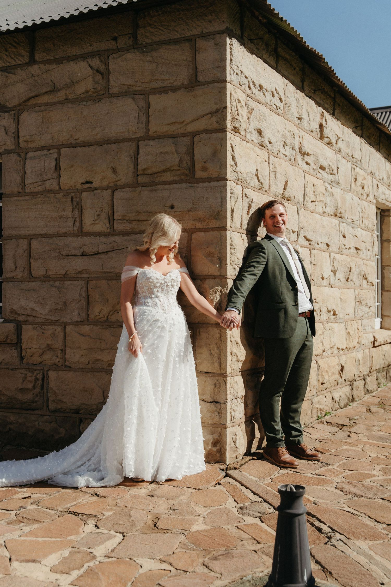 Bride in white lace gown and groom in green suit hold hands against a rustic stone wall on their wedding day.