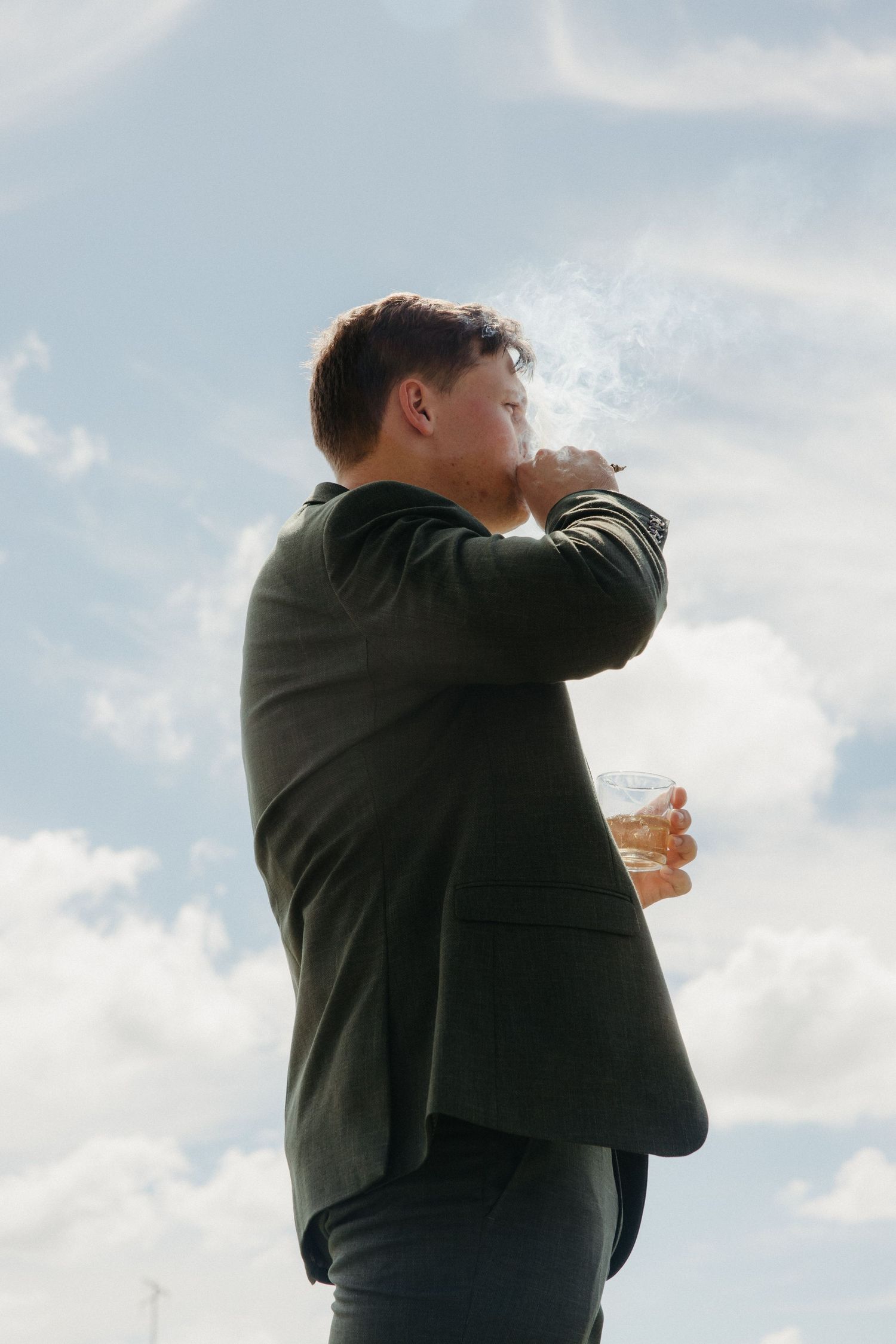 Man in dark suit smoking and holding whiskey glass outdoors against cloudy sky.