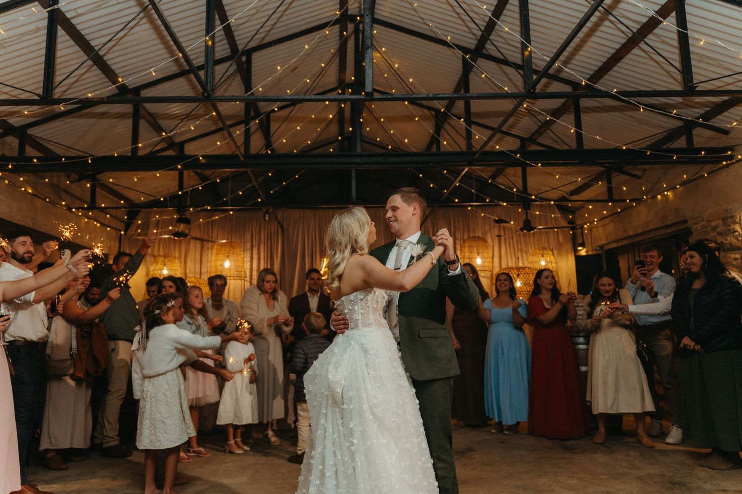 Bride and groom share first dance under fairy lights at rustic wedding venue surrounded by cheering guests.