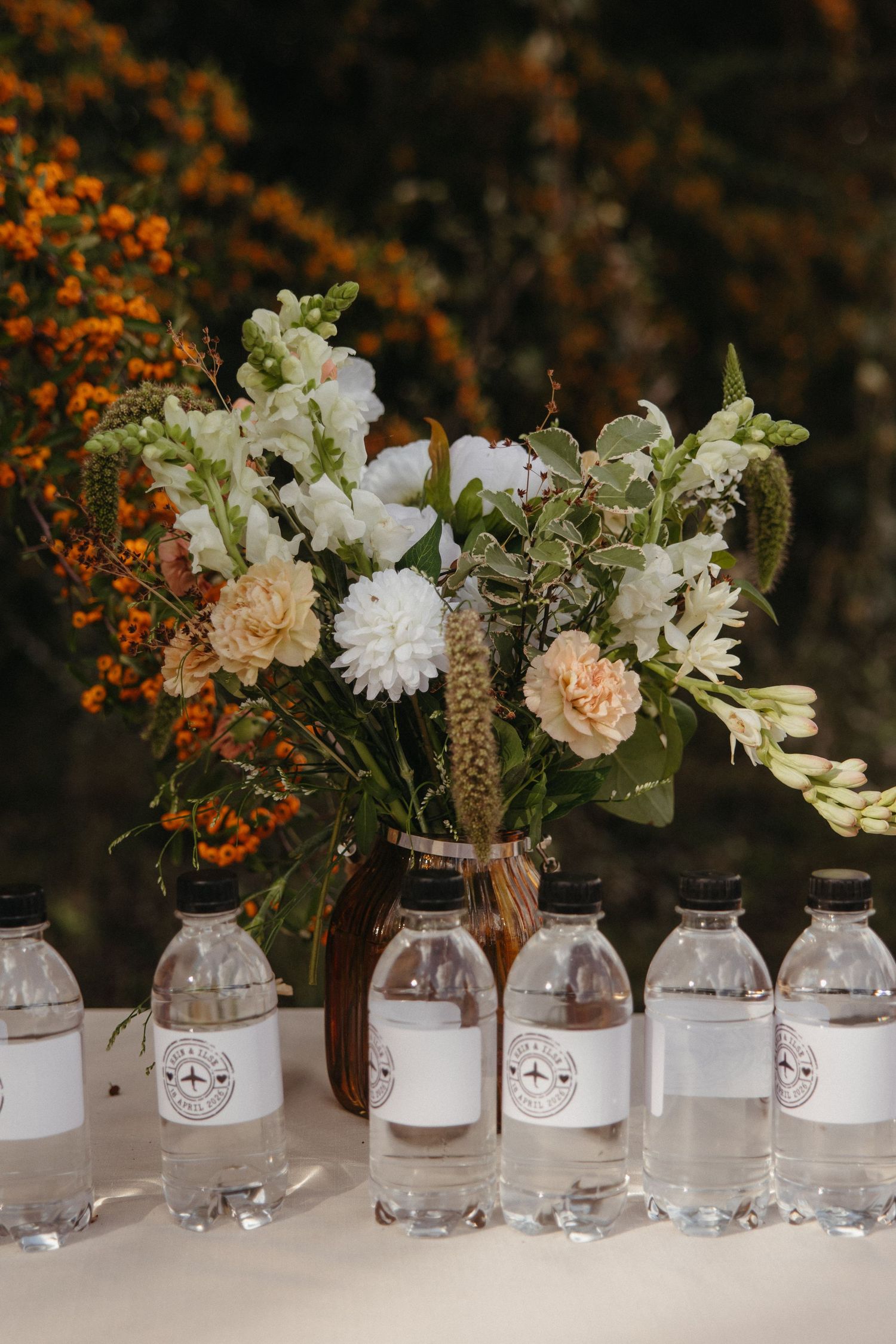 Elegant wedding table with labeled water bottles and a floral arrangement of white and peach blooms against autumn foliage.