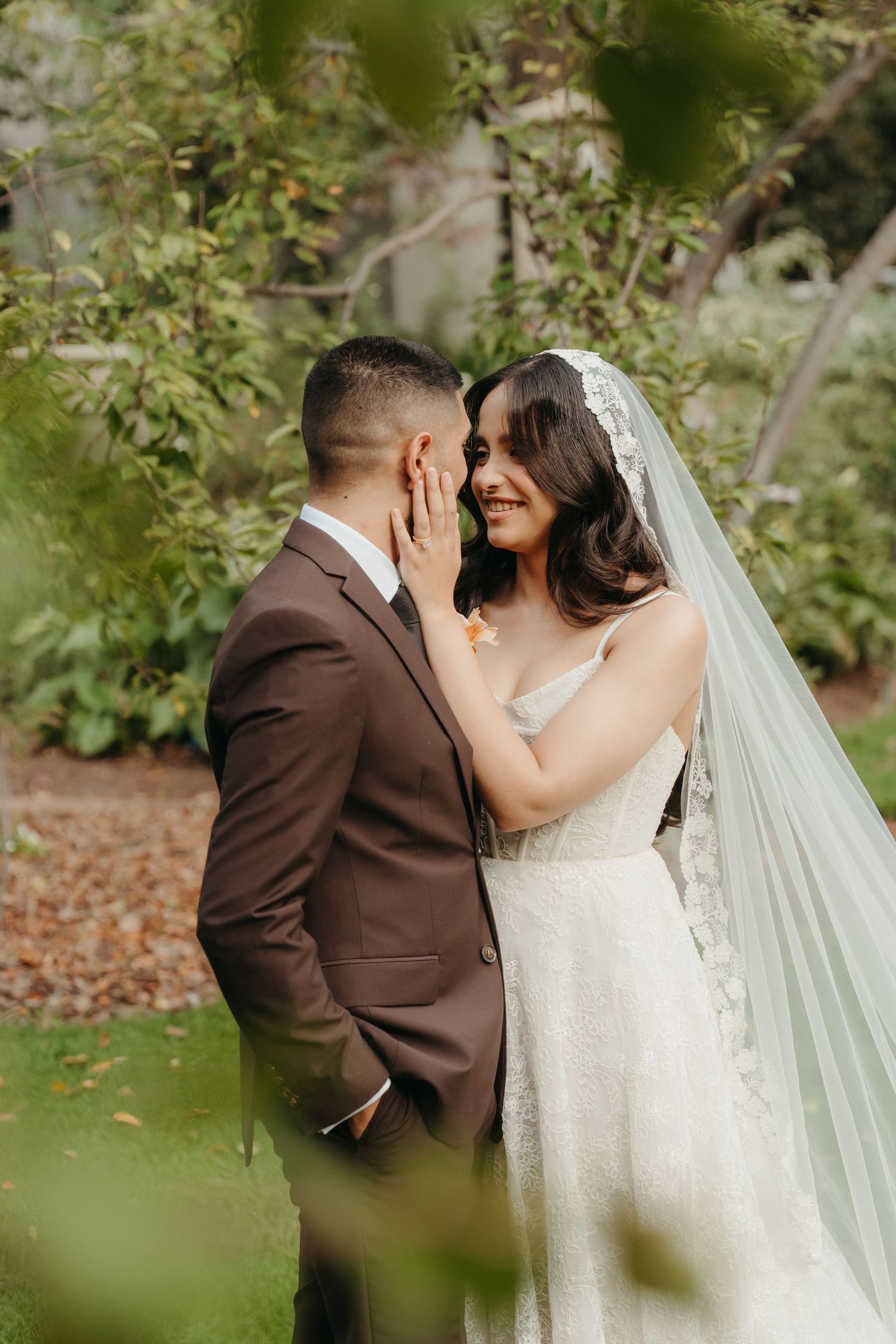 A bride in a lace gown and veil tenderly touches her groom's face as they share an intimate moment in a lush garden.