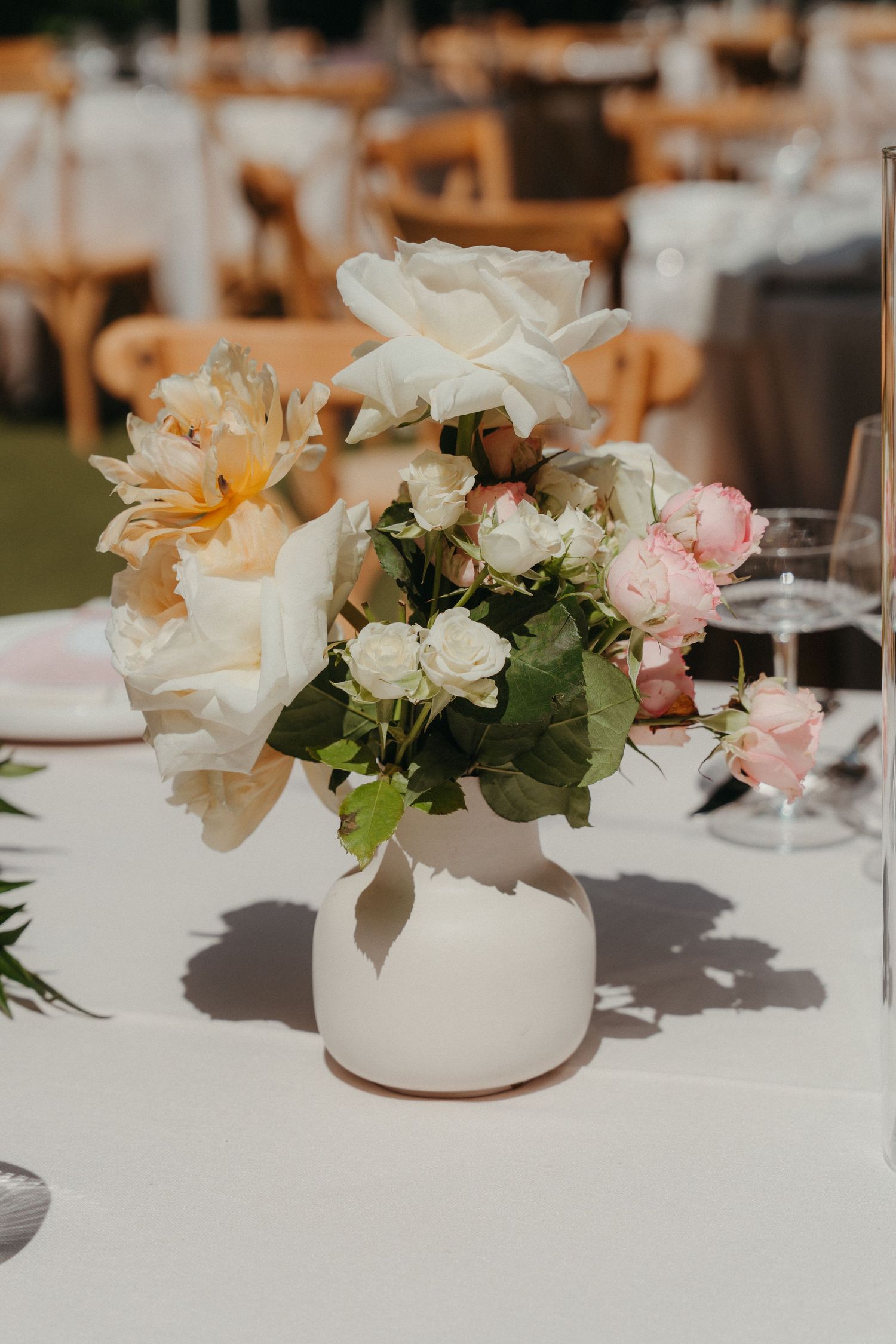 White ceramic vase with cream, blush pink roses and greenery centerpiece on white tablecloth at outdoor wedding reception.