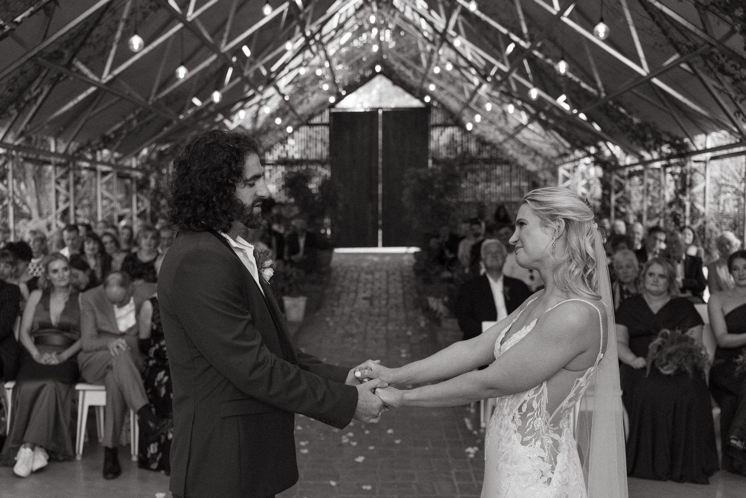Black and white photo of bride and groom holding hands at wedding ceremony in elegant greenhouse venue.