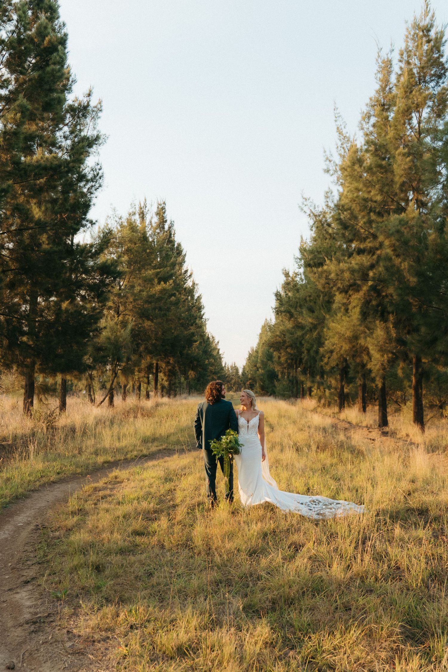 Bride and groom walking along a dirt path through tall pine trees in golden hour light, bride wearing a flowing white gown.