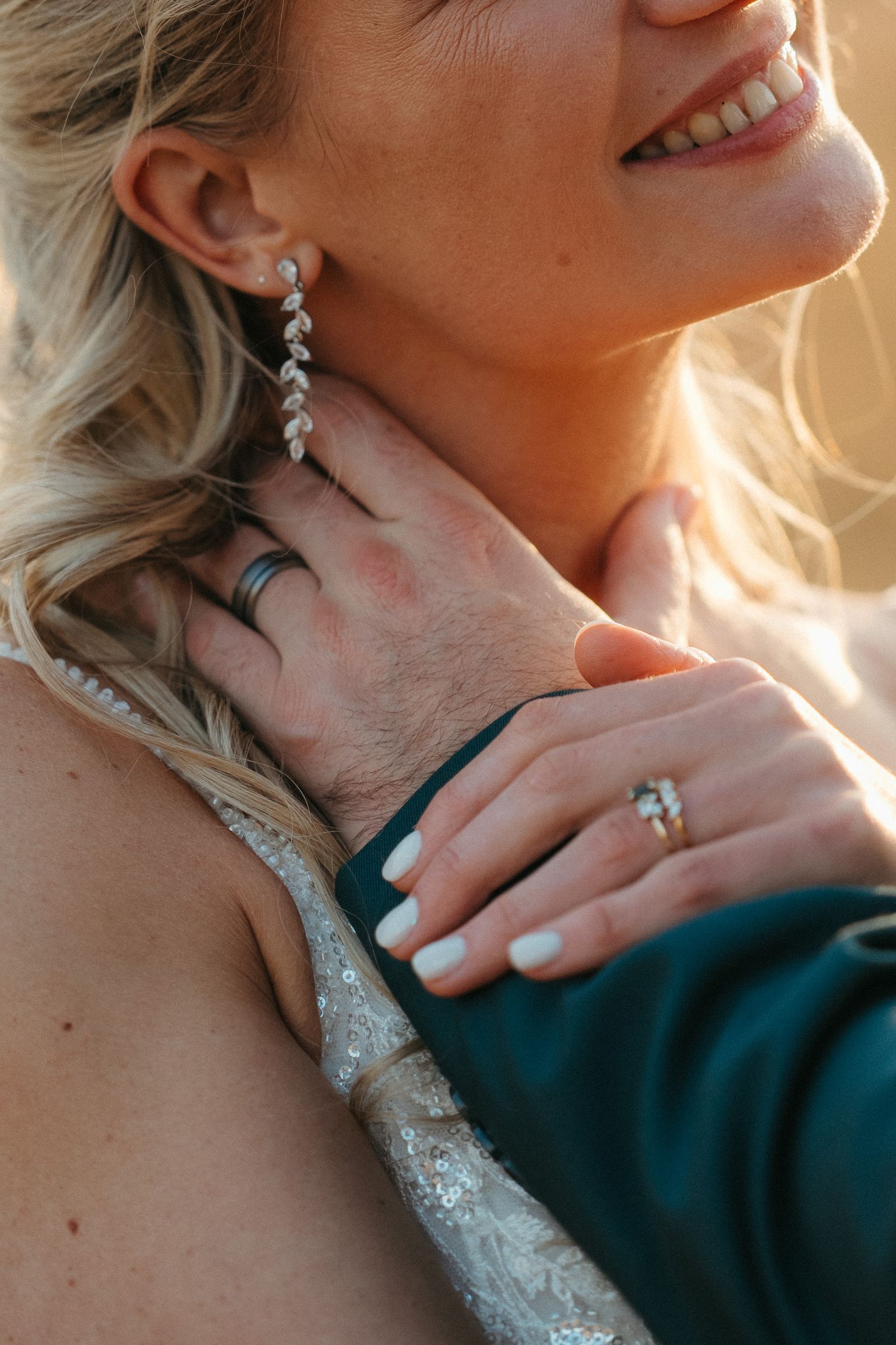 Close-up of bride smiling, wearing diamond drop earrings and engagement ring, with groom's hand on her neck.