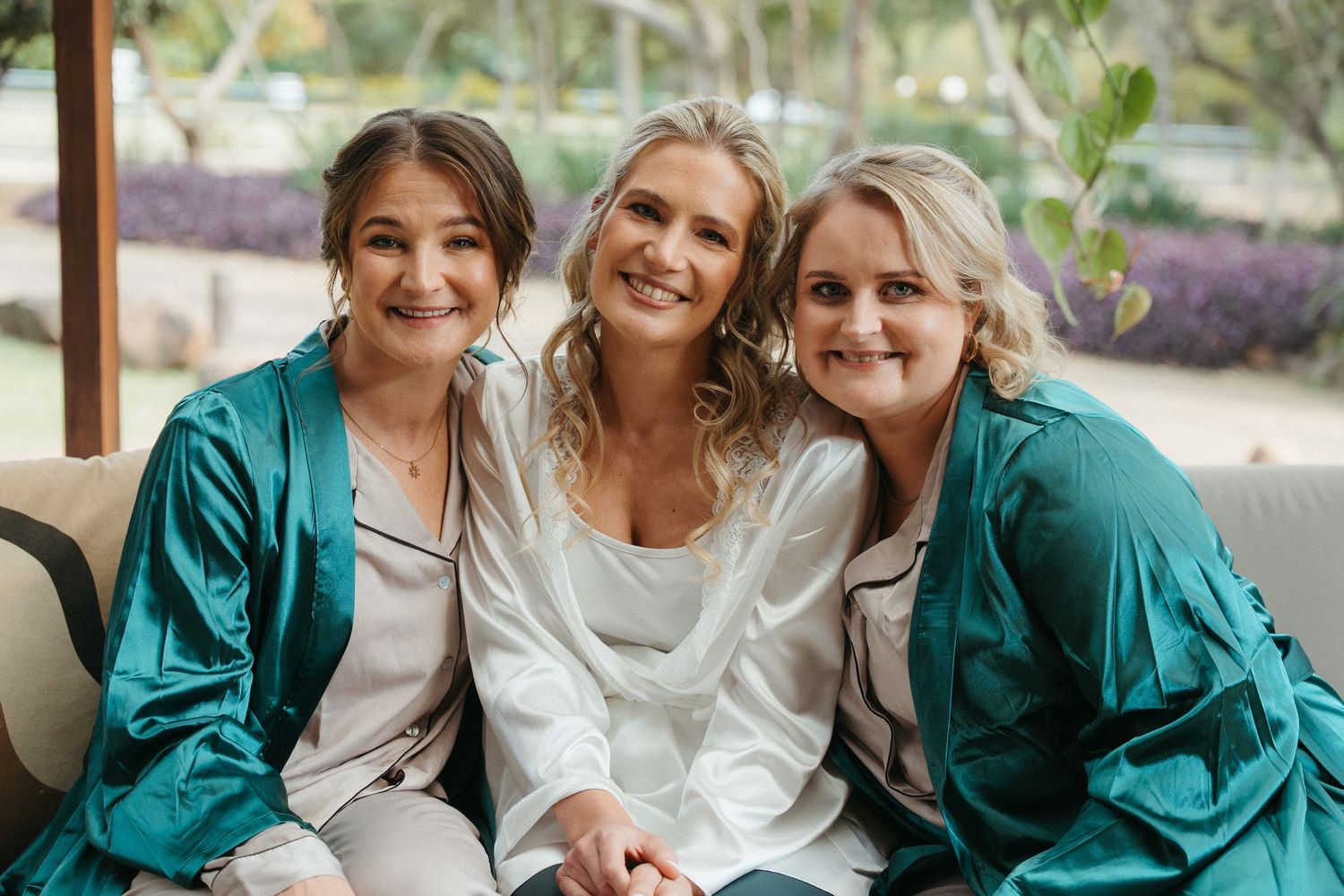 Three women in satin robes smile together on a couch, two in teal and one in white, likely a bridal party getting ready.