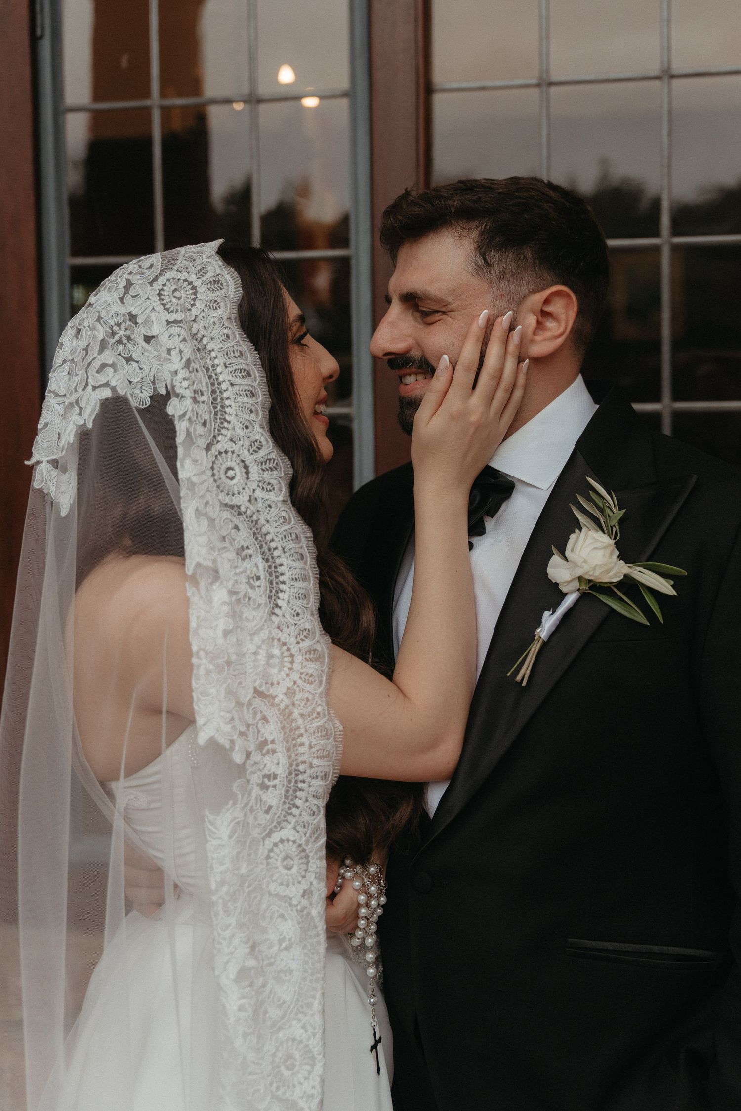 A bride in lace veil tenderly touches groom's face as they share a joyful moment in formal wedding attire.