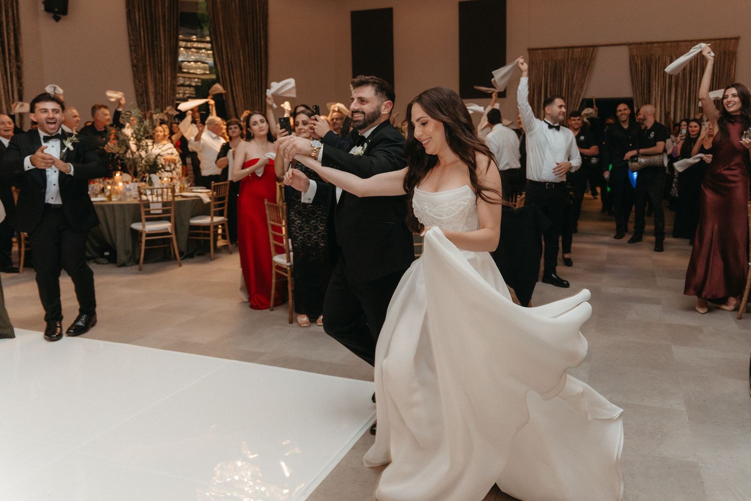 Bride and groom share first dance at wedding reception, bride twirling in white gown with guests celebrating in background.