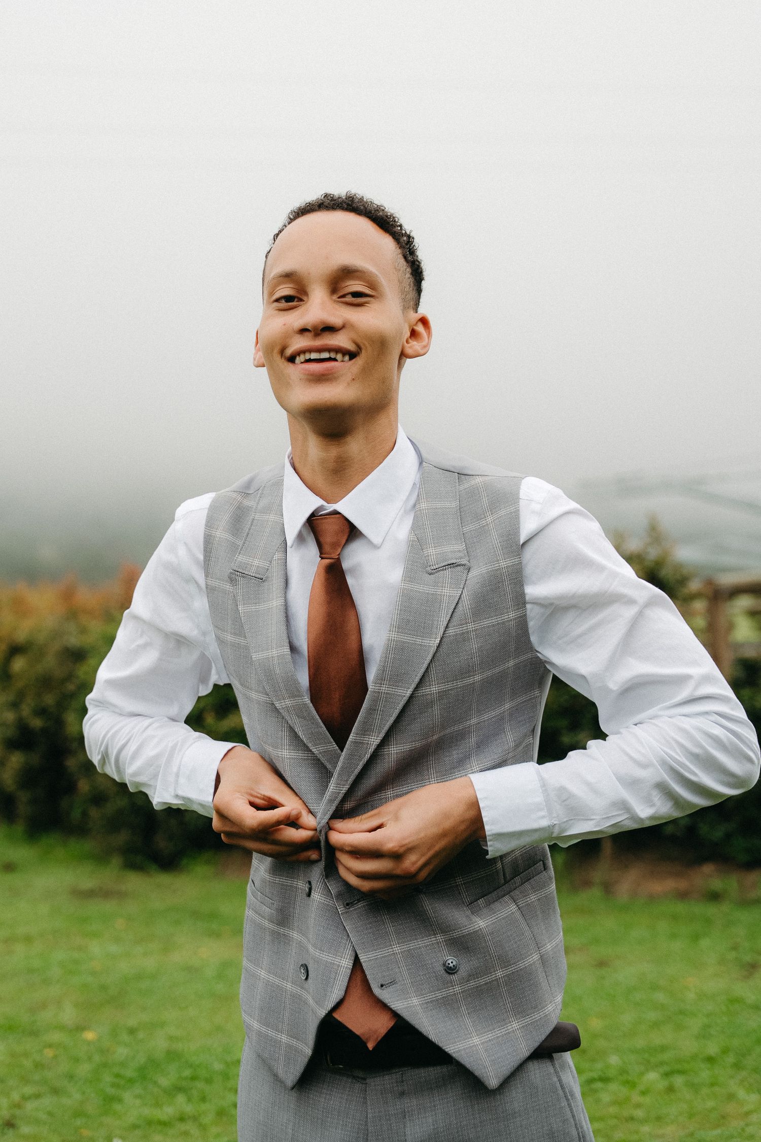 Smiling groom in gray plaid vest and brown tie buttoning his jacket outdoors on a foggy day in the Western Cape