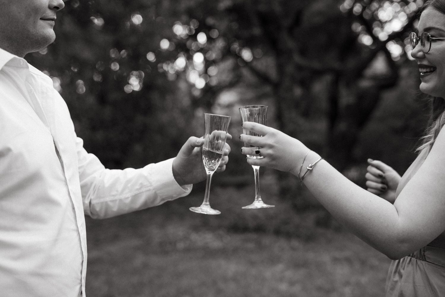 Two people toast with champagne glasses in black and white photography outdoors.