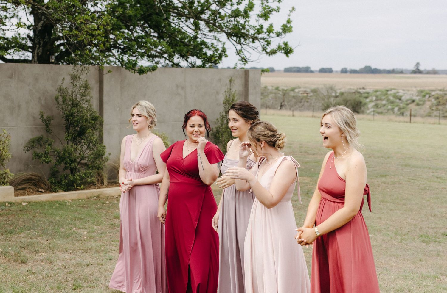 A group in elegant floor-length gowns in shades of pink and red share a joyful moment together outdoors.