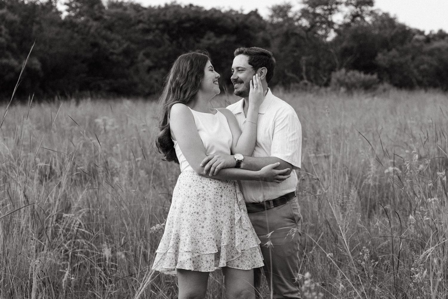 Black and white photo of a couple embracing and smiling in a field of tall grass.