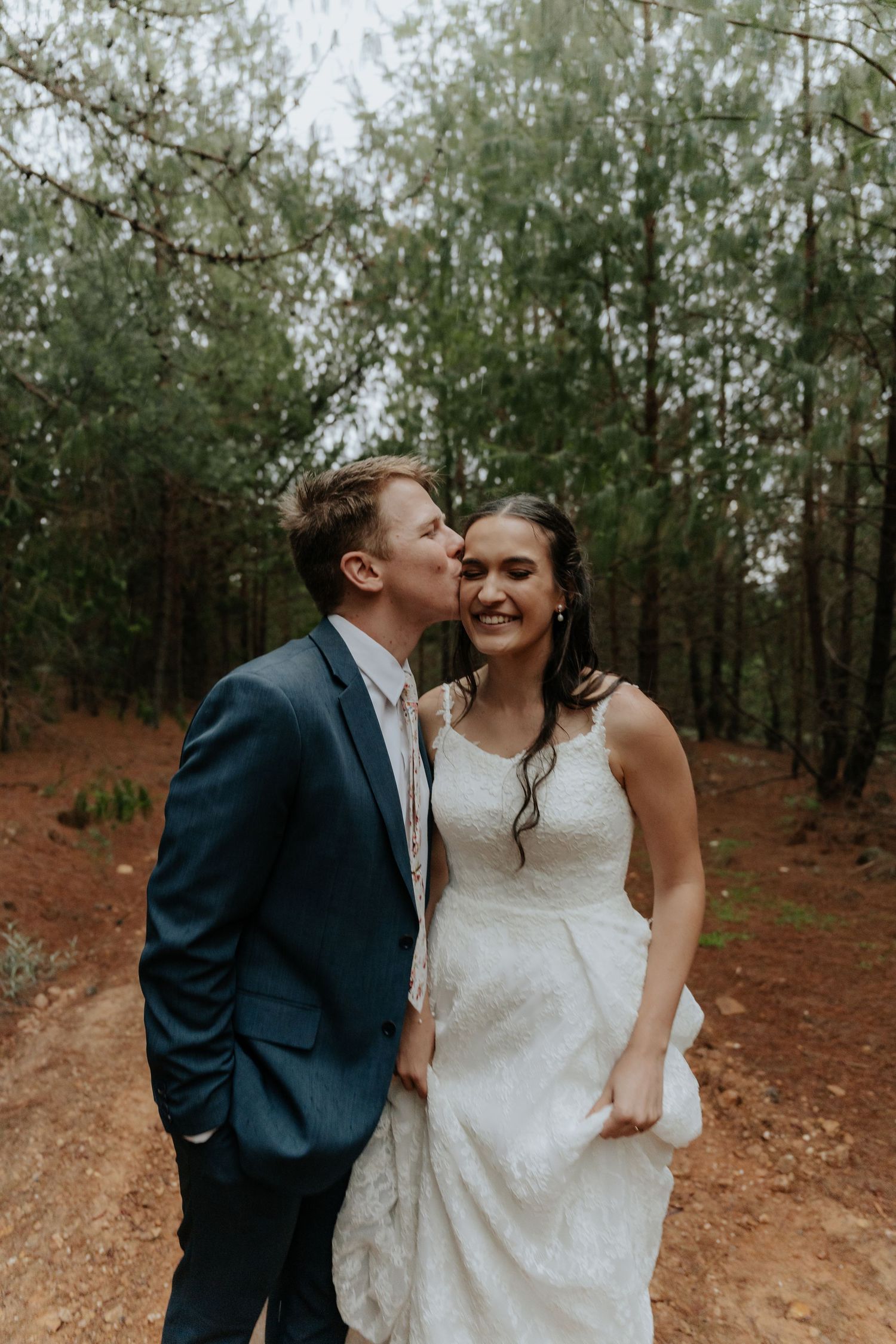 A romantic moment captured between a couple in formal attire standing among trees on a rustic path.