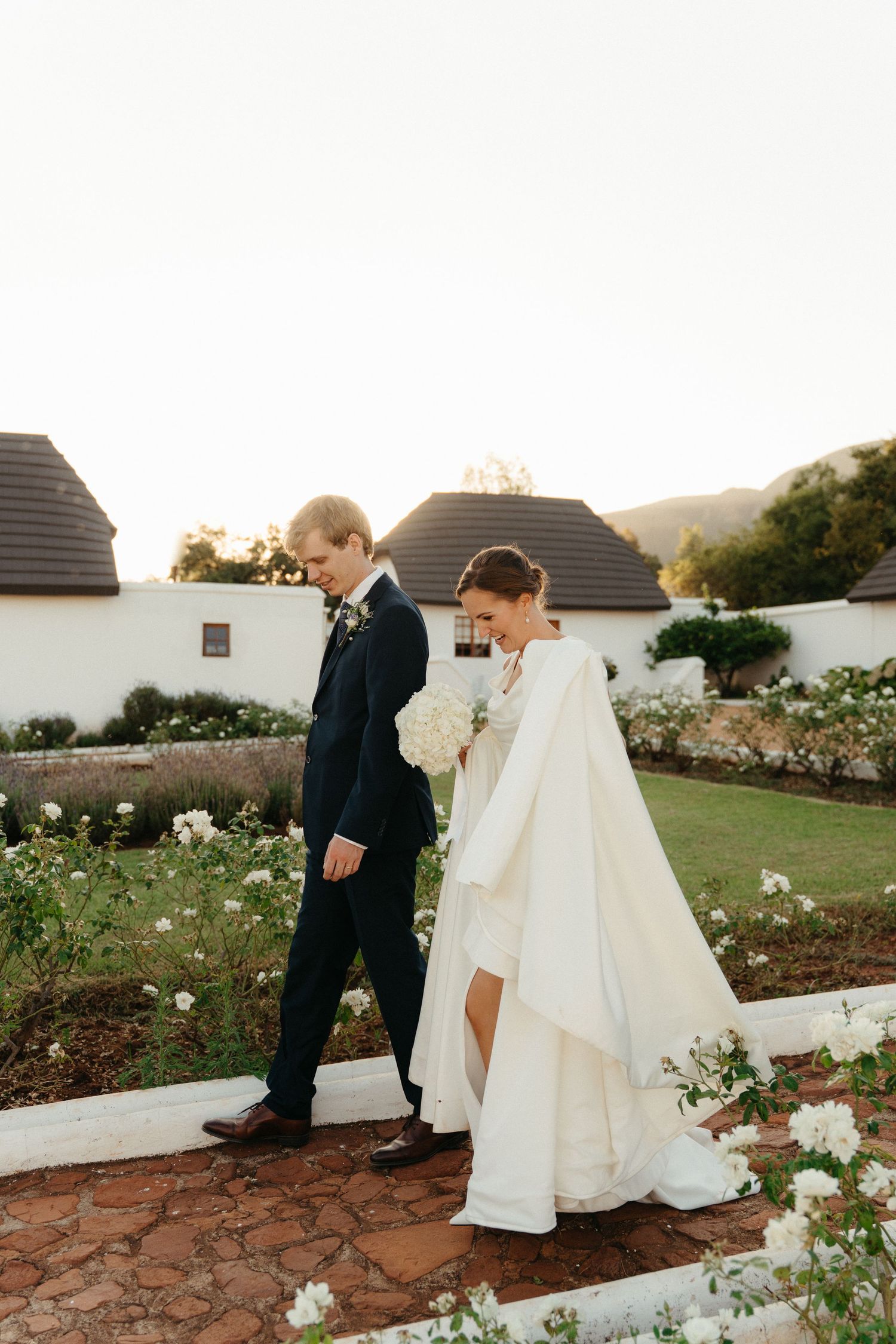 A couple walks together through a garden path lined with white roses on their wedding day at sunset.