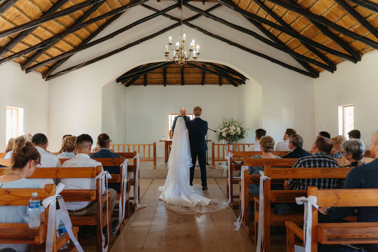 A small chapel wedding ceremony takes place in a rustic venue with wooden beams and pews under a chandelier.