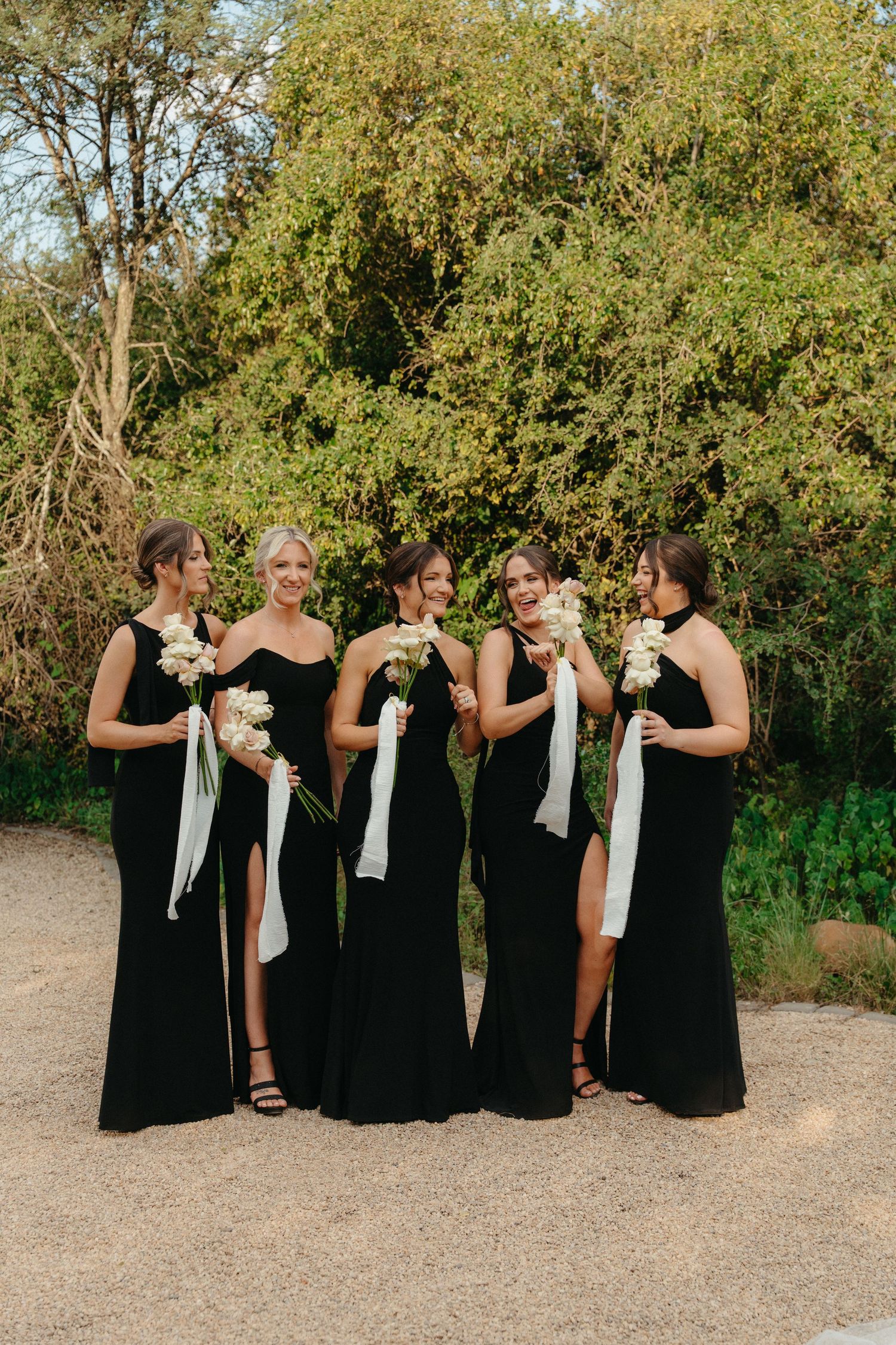 A group in black dresses laughs together while standing in a row on a dirt path surrounded by green trees.