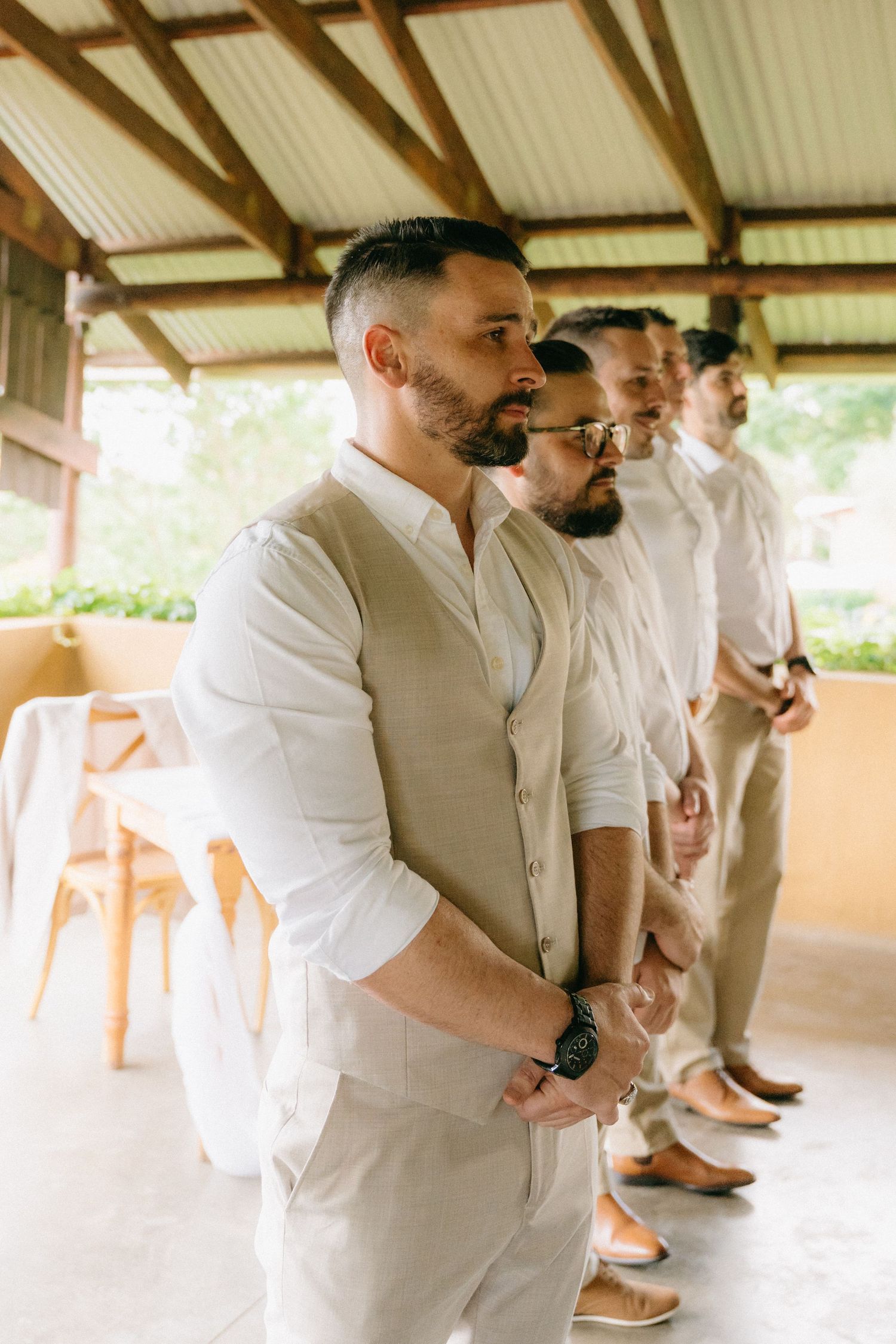 Groomsmen in light colored suits gather under covered outdoor area.
