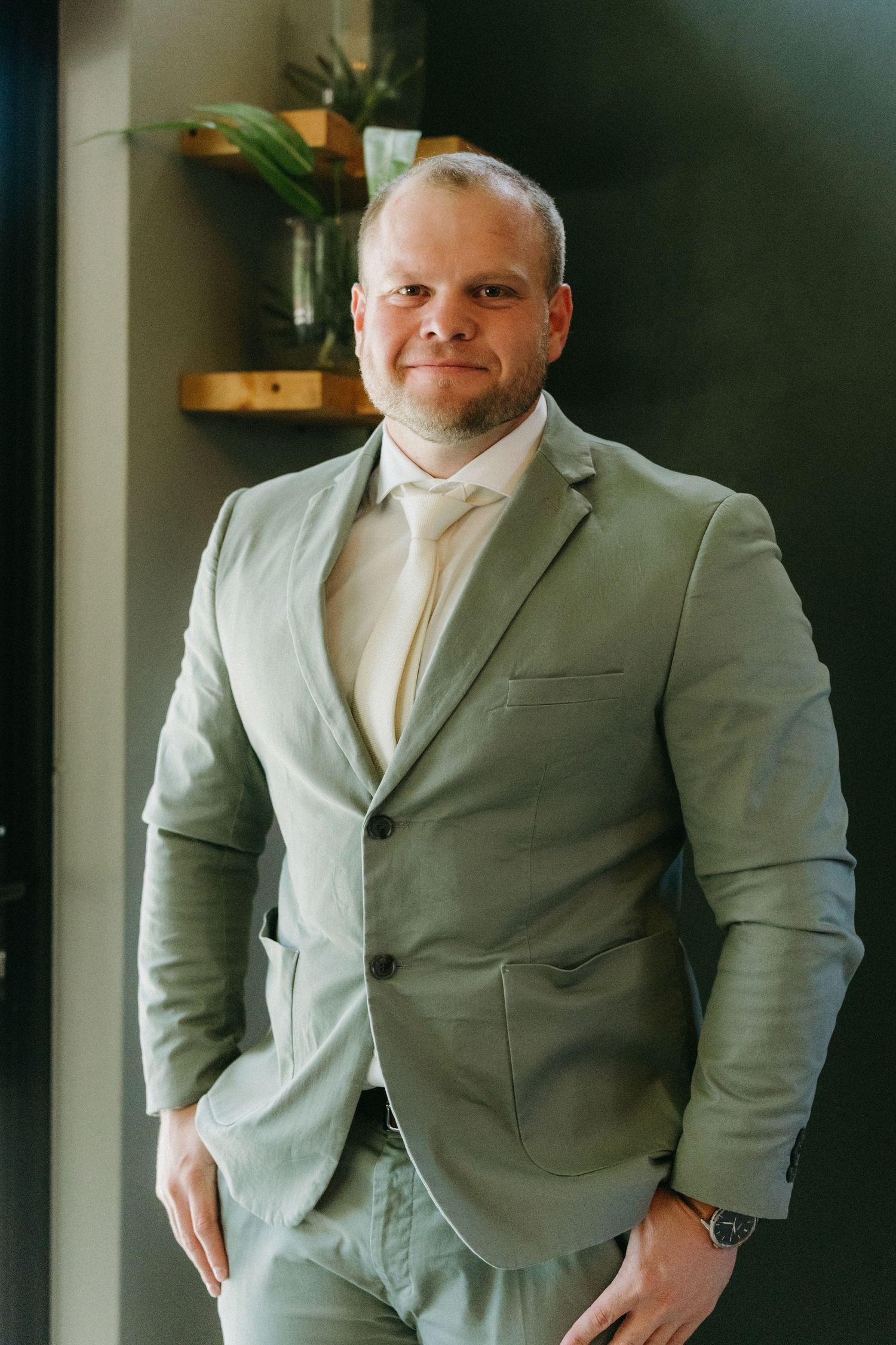 A person in a light gray suit poses confidently against a dark wall in an elegant indoor setting.