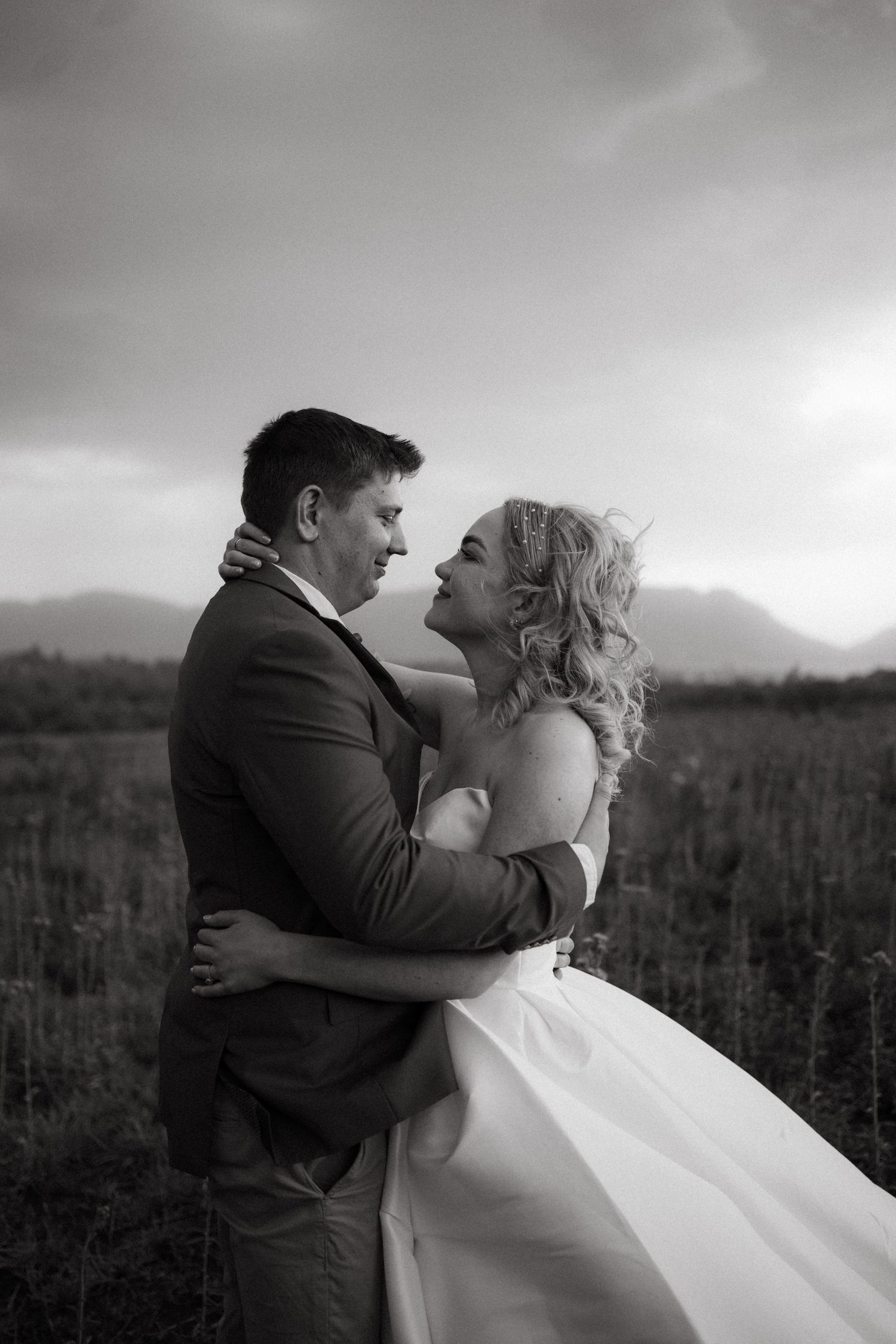 A romantic black and white wedding photo shows a couple embracing in a field with mountains in the background.