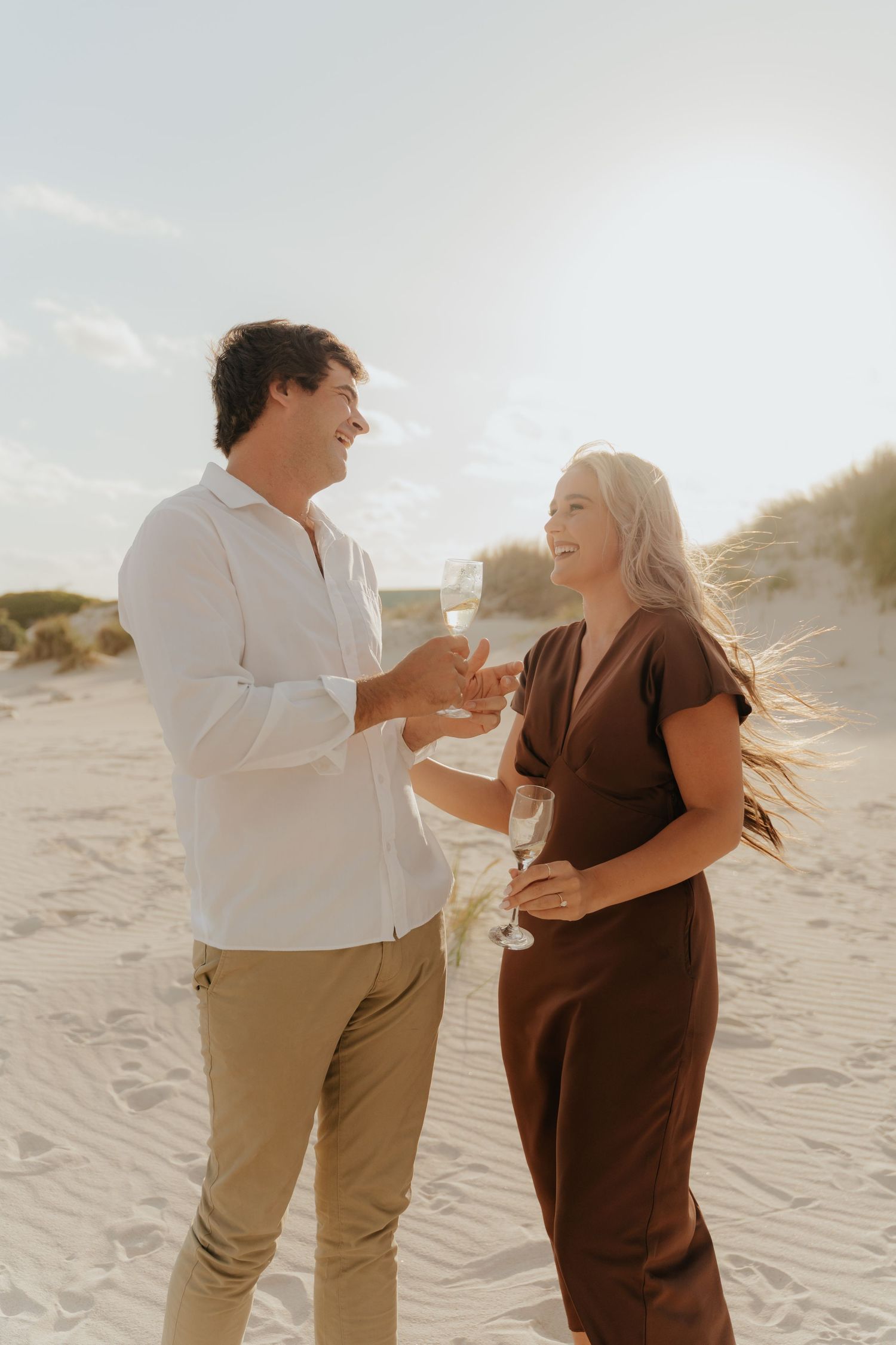 Happy couple toasting with champagne glasses on a sunny beach, dressed in white shirt and brown dress.