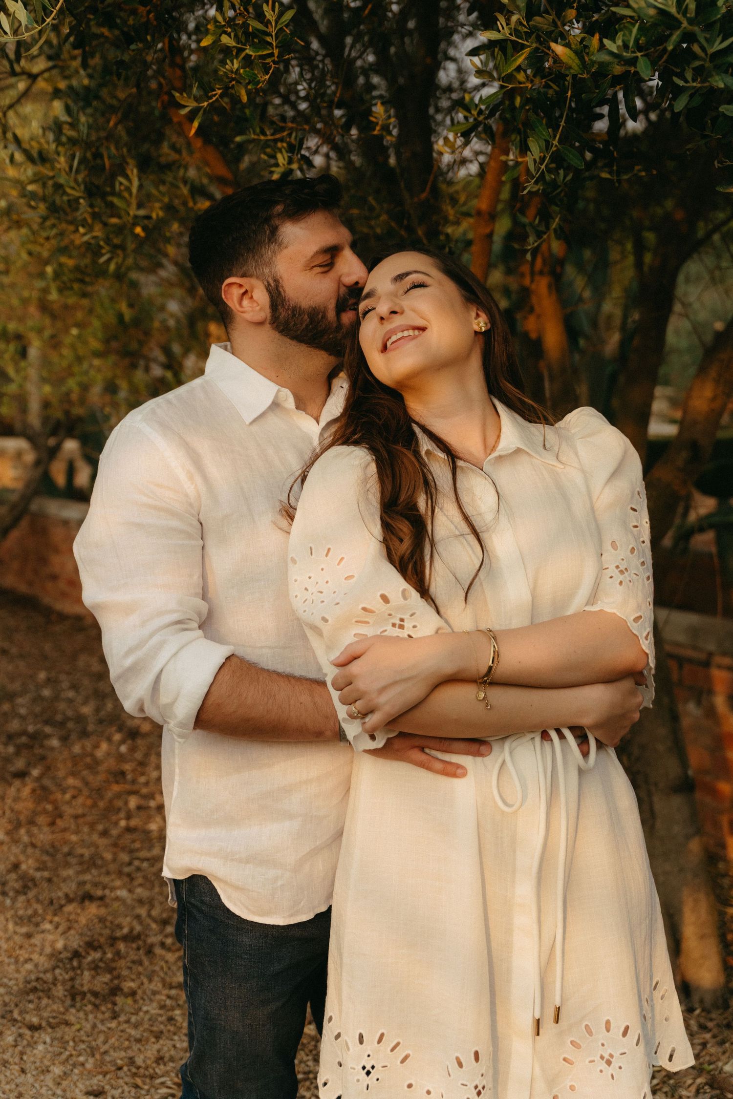 Romantic couple in white outfits embracing outdoors during golden hour among autumn trees.