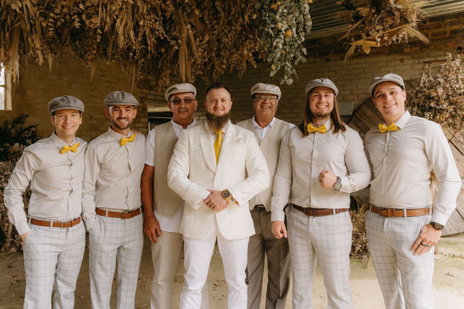 Groomsmen in matching white suits with yellow accents posing together in rustic barn setting.