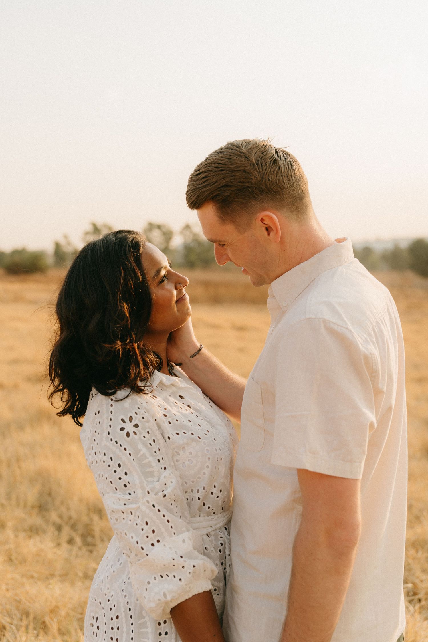 Romantic couple embraces in golden field during sunset photoshoot wearing white eyelet dress and light colored shirt.