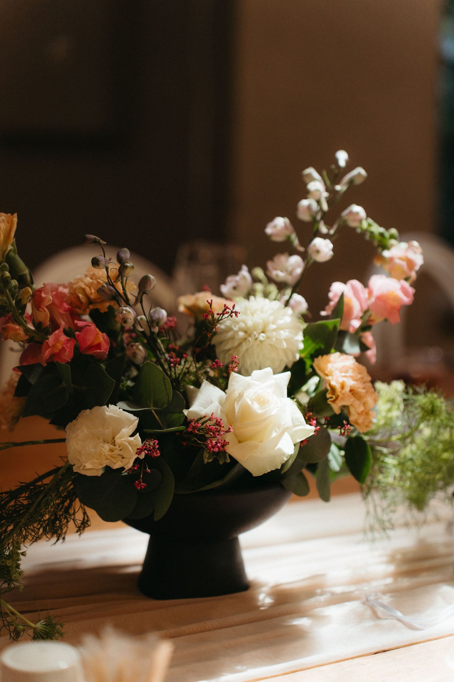 Wedding centerpiece arrangement featuring pink and white roses with delicate greenery on a long wooden table in warm lighting.