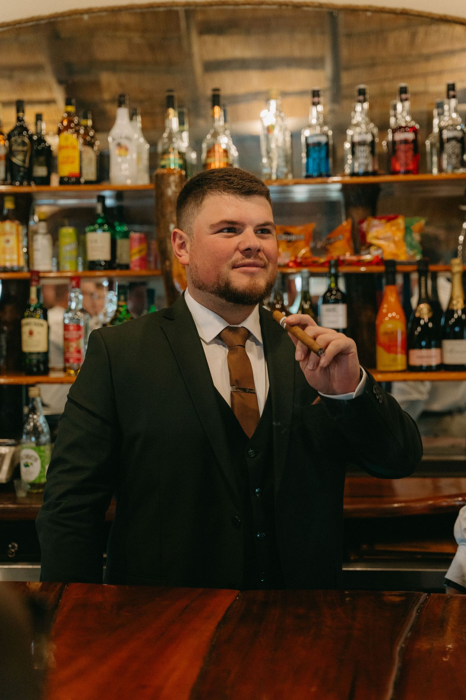 Well-dressed bartender in suit standing at wooden bar counter with shelves of liquor bottles.