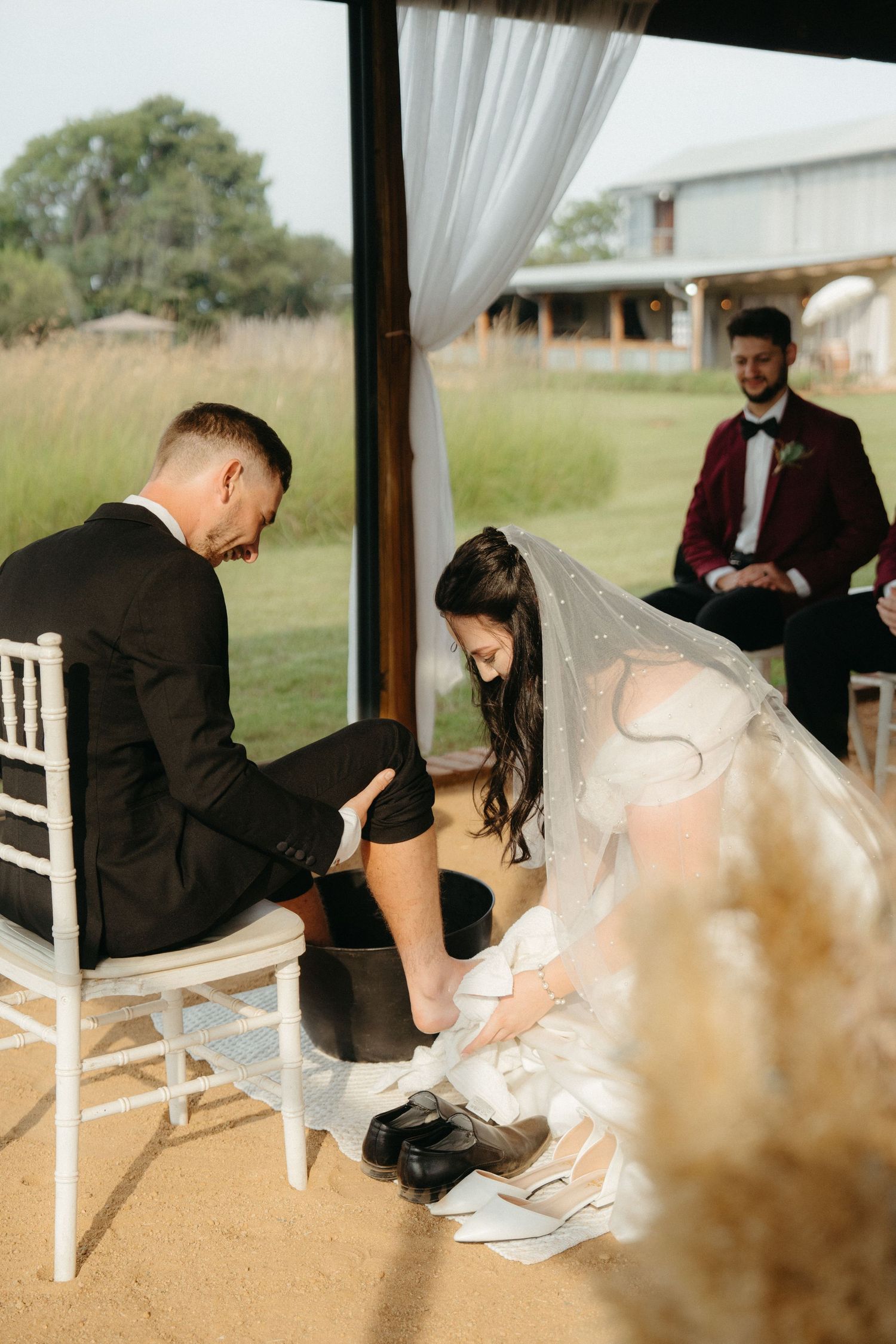 A touching wedding ceremony moment as a groom kneels to put on a shoe while a bride in white dress sits at an outdoor venue.