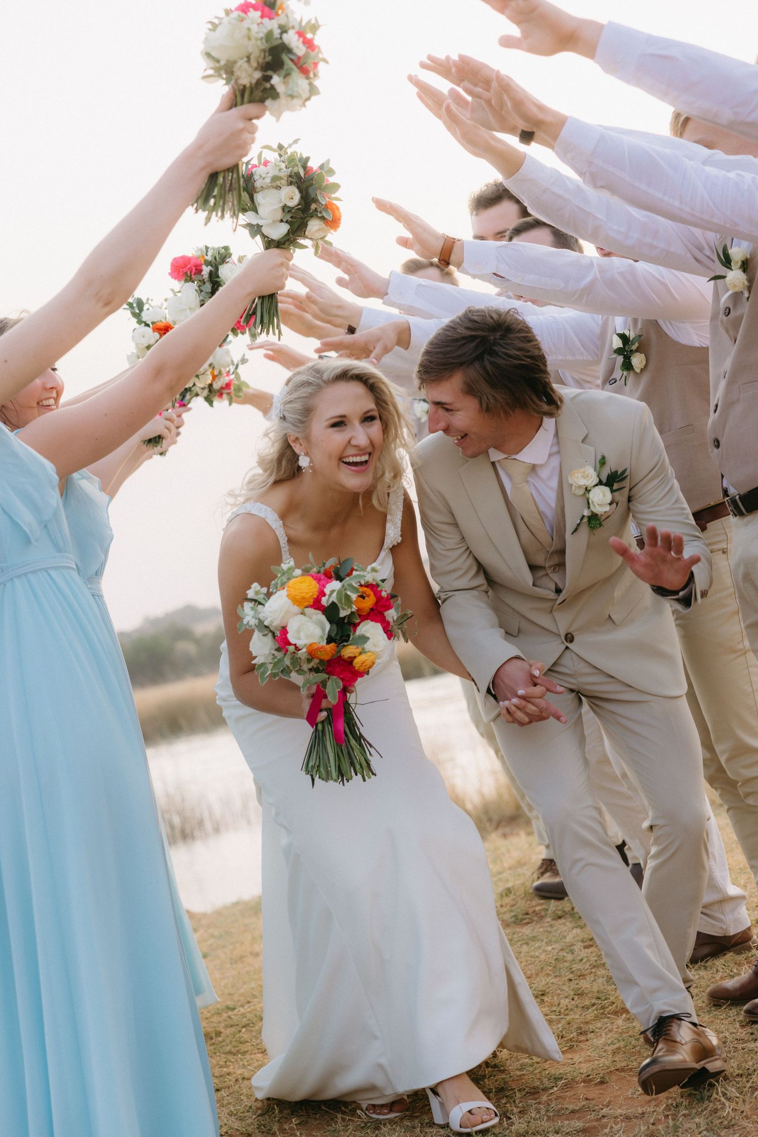 Wedding party forms a celebratory tunnel with raised arms in light blue and beige attire.