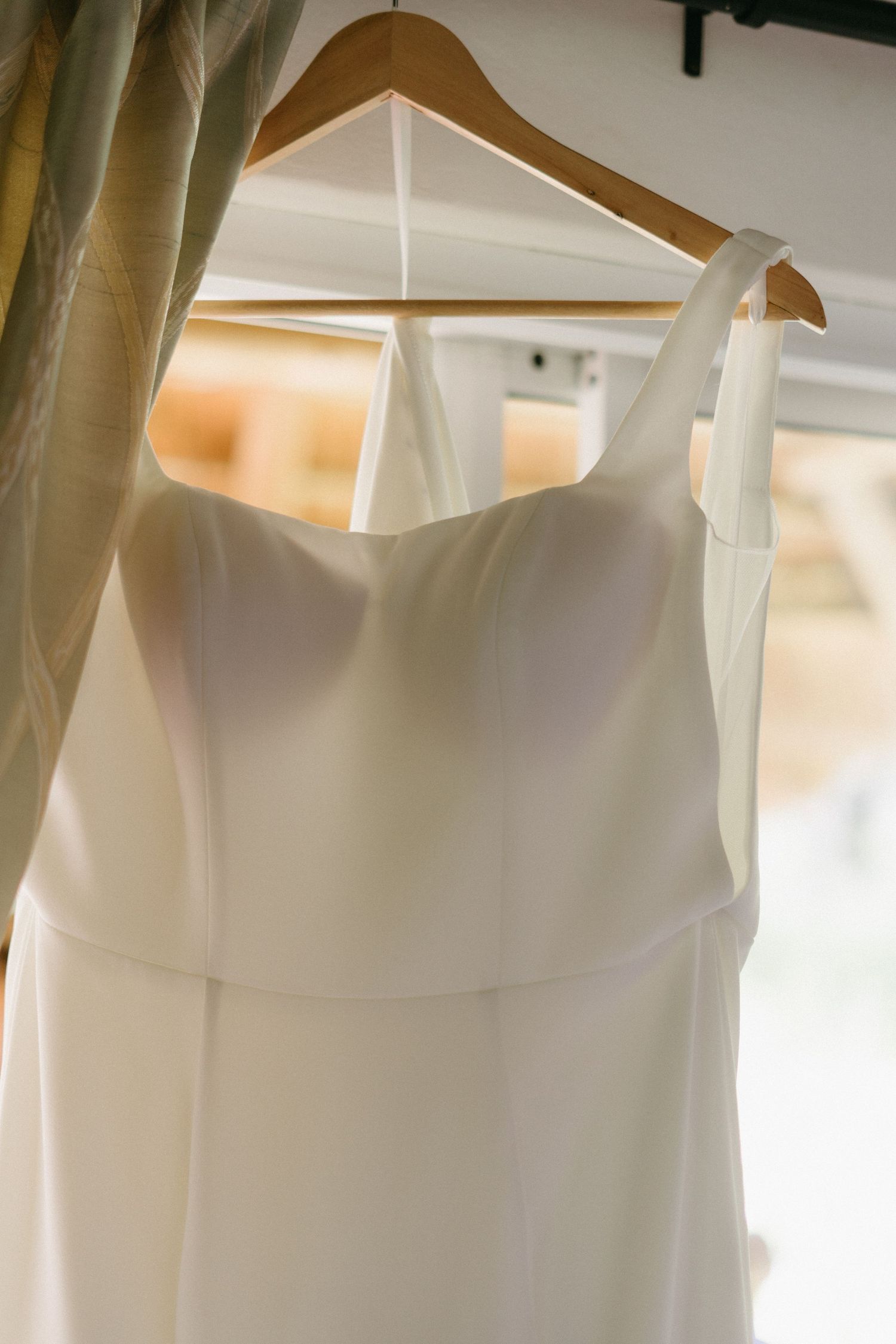 A white wedding dress hanging on a wooden hanger with soft natural lighting through curtains in the background.