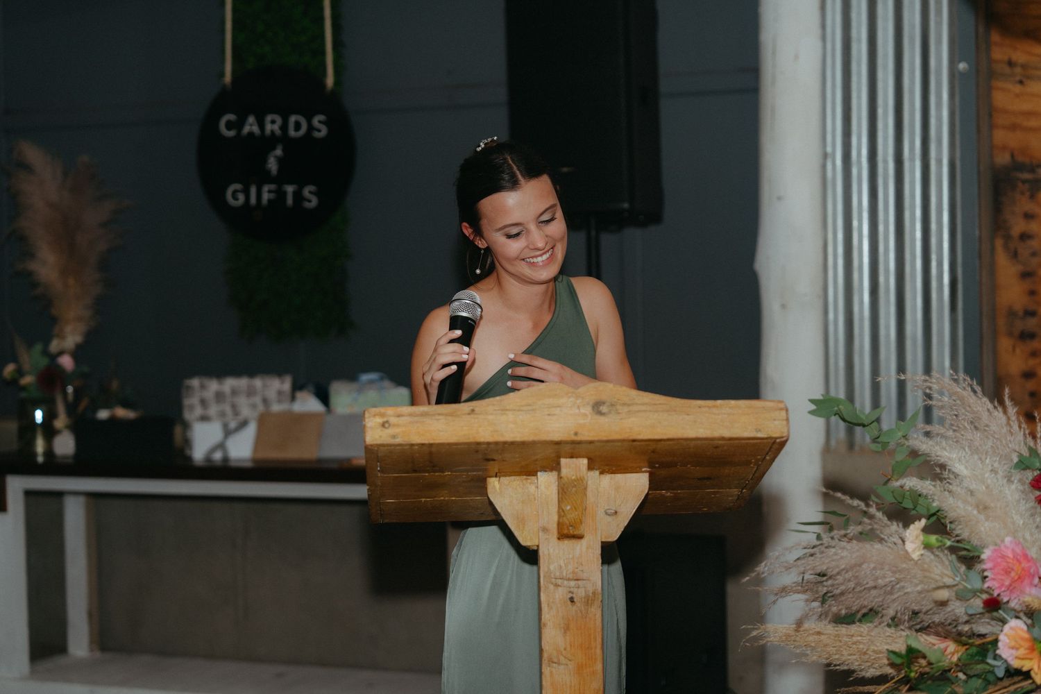 Bridesmaid in sage green dress reads speech from wooden podium during wedding reception.