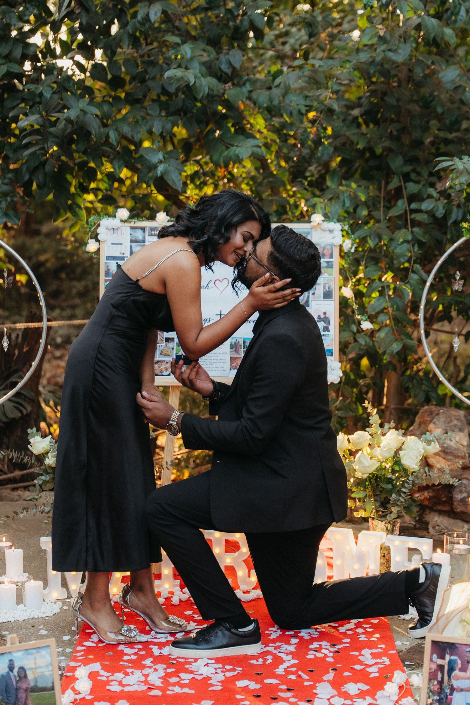 Couple in black outfits sharing a kiss during a romantic outdoor proposal with candles and floral decor.