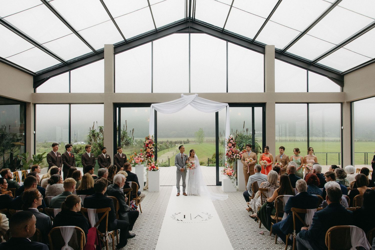 Elegant indoor wedding ceremony in a glass greenhouse venue with floral arch, white aisle runner, and lush garden views.