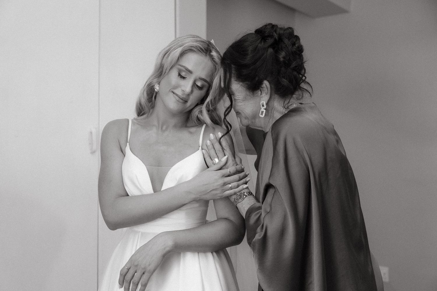Black and white photo of a bride in a white gown sharing an emotional embrace with her mom in a satin dress before the wedding.