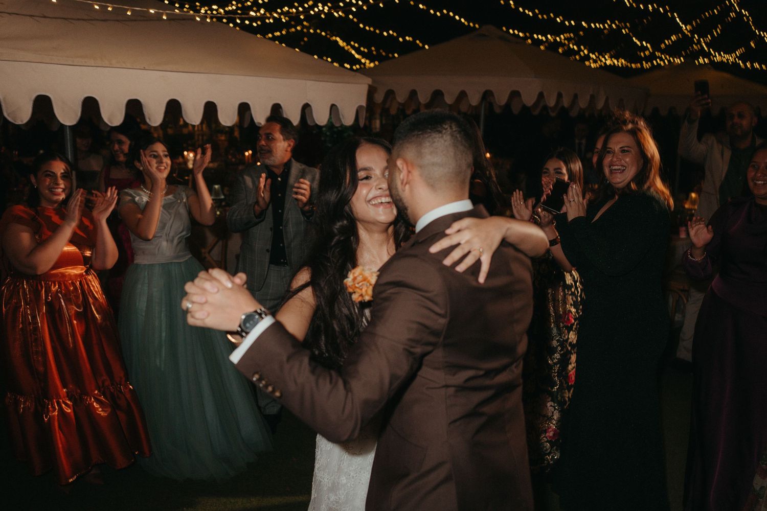 Bride and groom share their first dance surrounded by cheering guests at a romantic wedding reception with fairy lights.