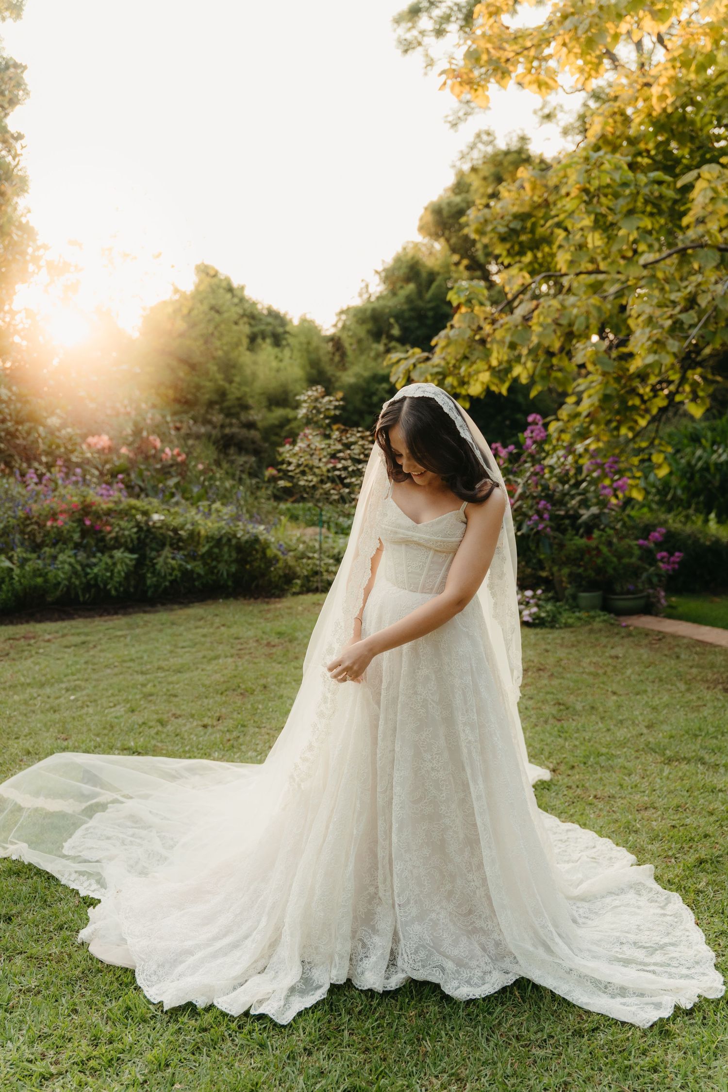 Bride in lace wedding gown with veil stands in sunlit garden, looking down at her dress amid blooming flowers.