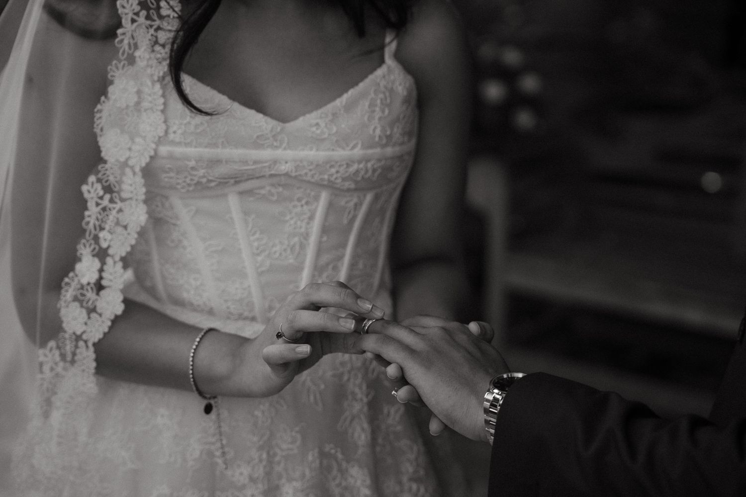 Black and white wedding photo of bride in lace dress receiving ring from groom during ceremony.