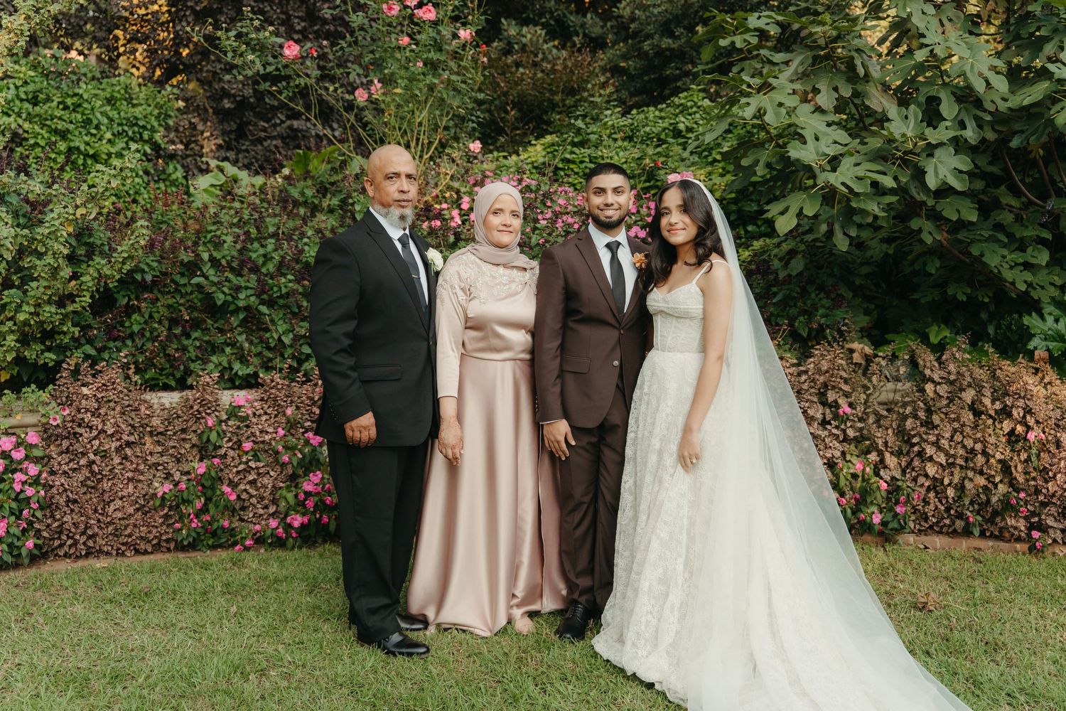 Wedding party of four posing outdoors, bride in white gown with veil, groom in brown suit, with elegantly dressed parents.