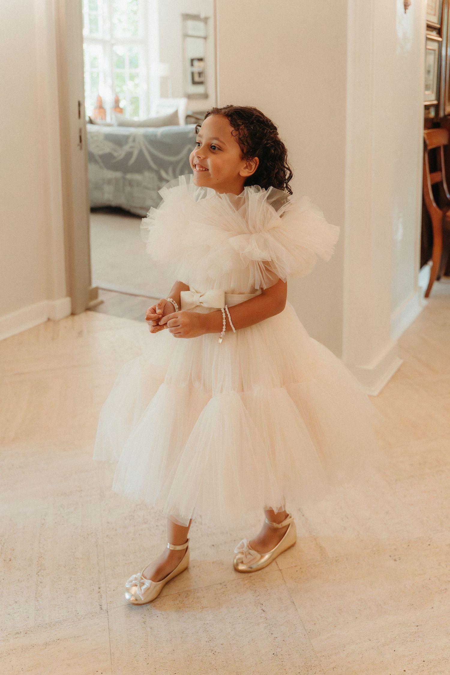 Young girl in white tulle flower girl dress with ruffled shoulders and gold flats, smiling indoors.